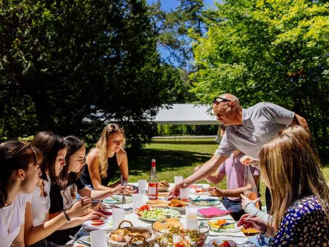 L'image capture un moment convivial d'un déjeuner en plein air, où un groupe de personnes partage un repas autour d'une table bien garnie dans un cadre verdoyant et ensoleillé.