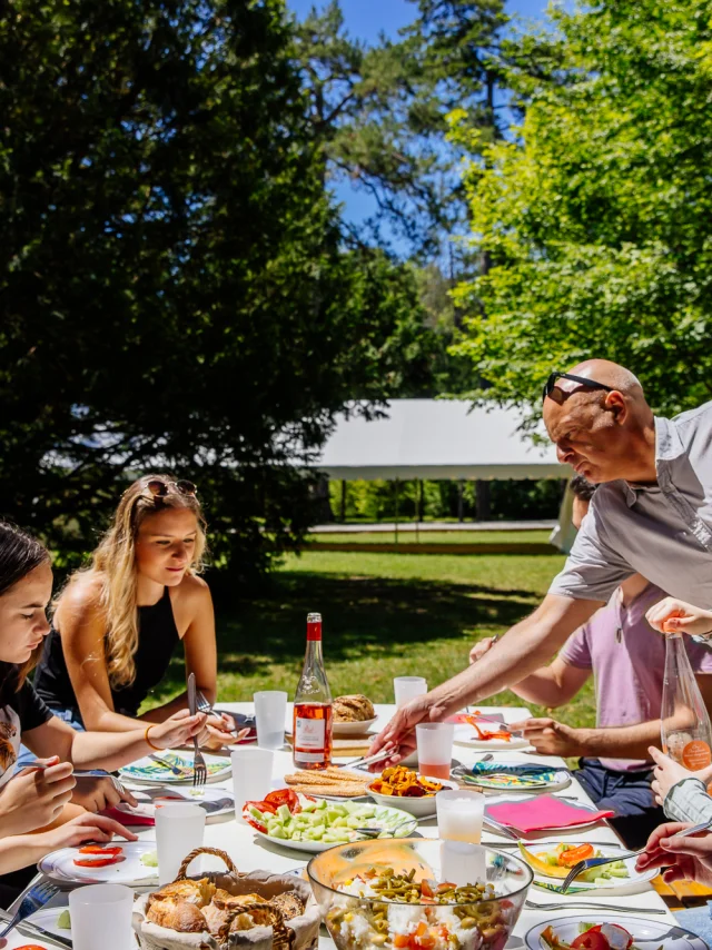 L'image capture un moment convivial d'un déjeuner en plein air, où un groupe de personnes partage un repas autour d'une table bien garnie dans un cadre verdoyant et ensoleillé.