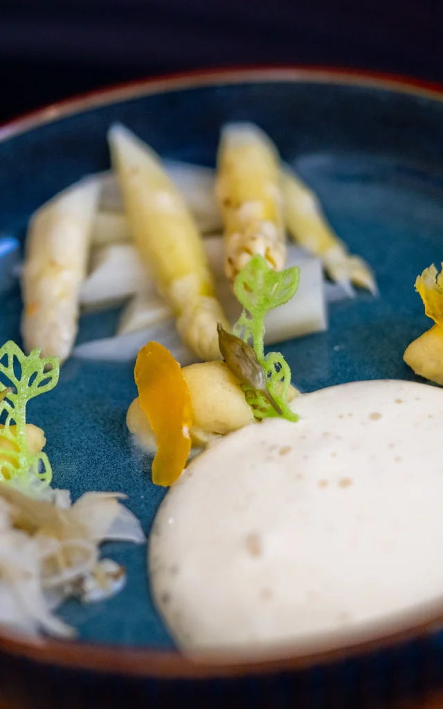 Assiette bleue contenant une préparation gastronomique à base d’asperges blanches, accompagnée de mousse onctueuse, de purée, de légumes finement découpés et de décorations vertes en forme de feuilles.
