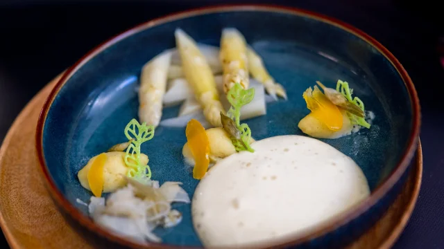Assiette bleue contenant une préparation gastronomique à base d’asperges blanches, accompagnée de mousse onctueuse, de purée, de légumes finement découpés et de décorations vertes en forme de feuilles.