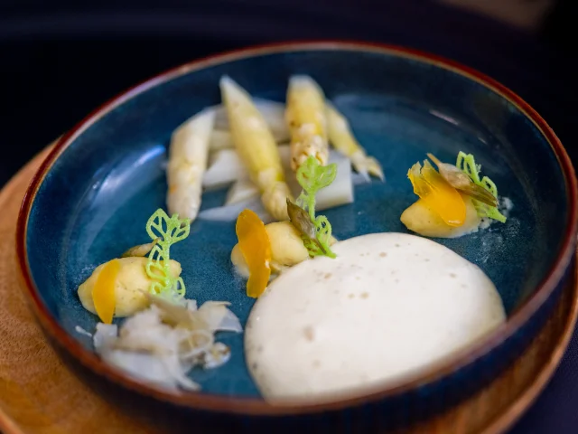 Assiette bleue contenant une préparation gastronomique à base d’asperges blanches, accompagnée de mousse onctueuse, de purée, de légumes finement découpés et de décorations vertes en forme de feuilles.
