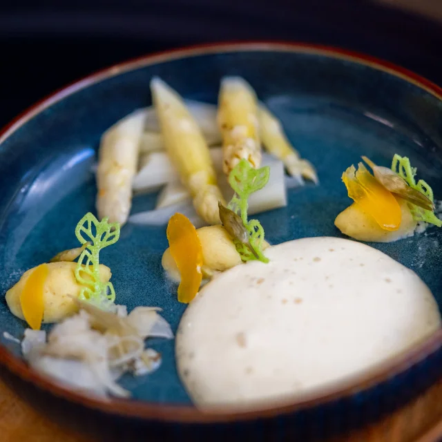 Assiette bleue contenant une préparation gastronomique à base d’asperges blanches, accompagnée de mousse onctueuse, de purée, de légumes finement découpés et de décorations vertes en forme de feuilles.