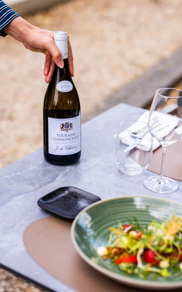 Main tenant une bouteille de vin blanc Touraine Chenonceaux millésime 2023 sur une table en extérieur, avec une assiette de salade fraîche et deux verres à vin posés à côté.