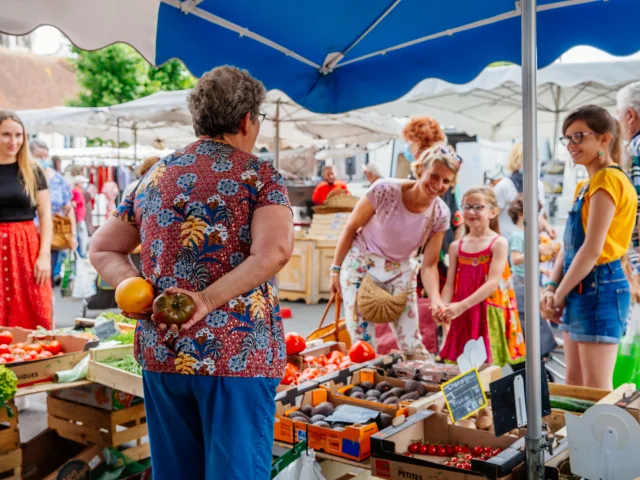 Une scène vivante dans un marché en plein air où une femme de dos tient deux tomates anciennes pendant qu'une autre femme et des enfants sourient devant un étal rempli de légumes frais.