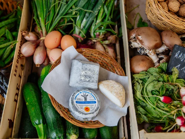 Un panier de fromages de chèvre au centre d’un étal coloré avec des courgettes, œufs, champignons, radis, oignons et autres légumes de saison, typique d’un marché local.