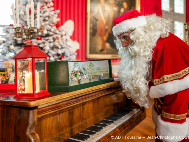 Père Noël observant une scène miniature posée sur un piano décoré pour Noël au Château Royal d’Amboise.