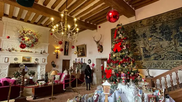 Grande salle du Château Gaillard décorée pour Noël avec un grand sapin illuminé, des miniatures hivernales, des décorations suspendues et une cheminée ornée.