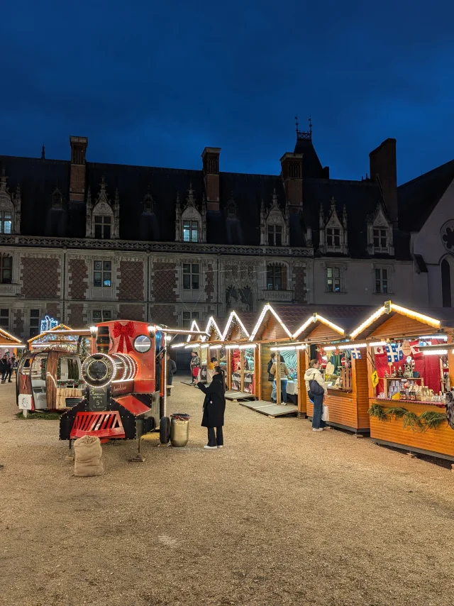 Marché de Noël illuminé devant le Château Royal de Blois, avec chalets en bois et décorations festives à la tombée de la nuit.