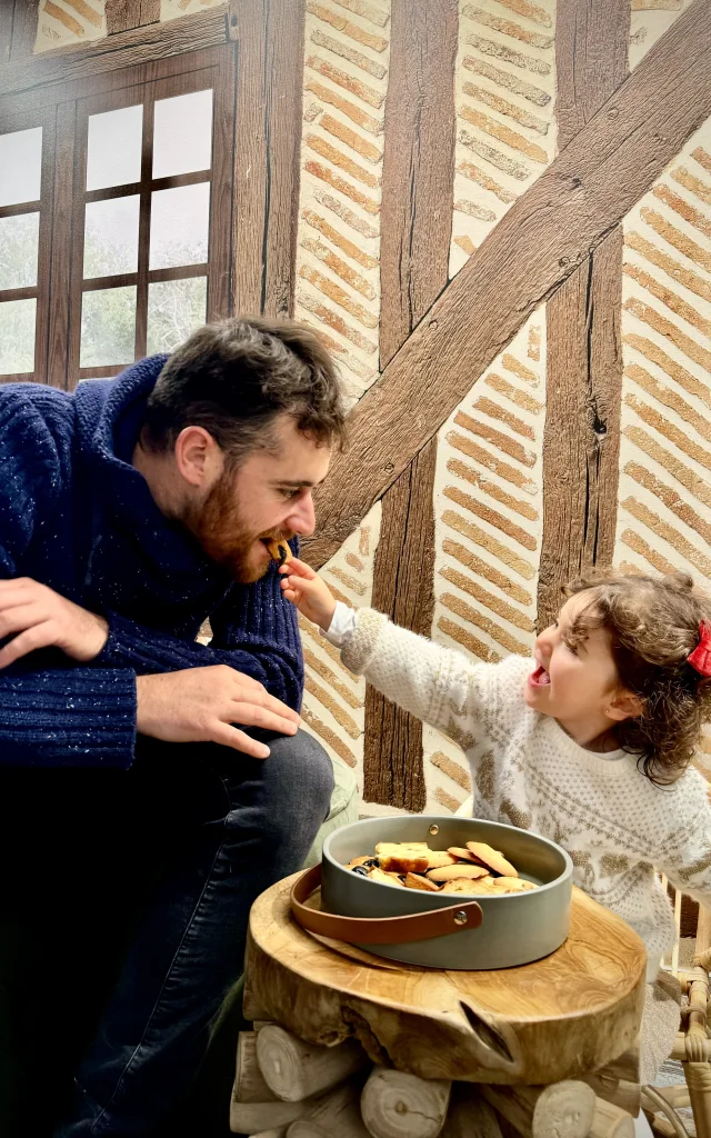 Un homme barbe en pull bleu marine et une petite fille bouclée en pull blanc crème jouent ensemble dans une pièce à colombages. La fillette tend un biscuit à l'homme, qui fait mine de le croquer, tous deux souriants et complices. Une boîte de biscuits repose sur une petite table en bois devant eux.