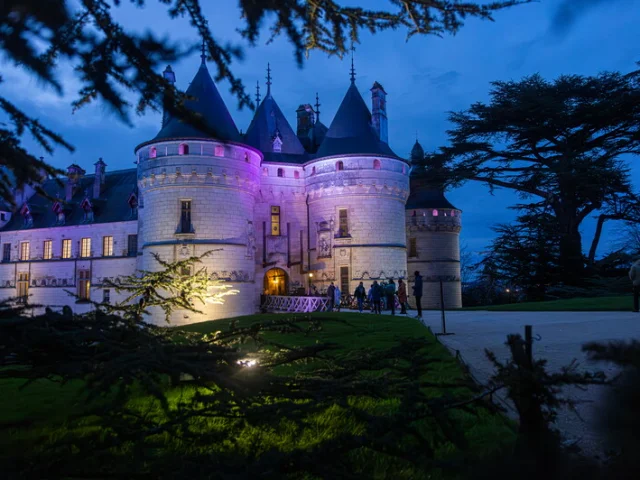 Façade du Château de Chaumont-sur-Loire illuminée en violet et bleu à la tombée de la nuit, avec des visiteurs entrant dans la cour.