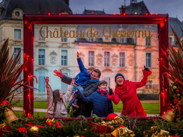 Une famille pose joyeusement dans un grand cadre décoratif rouge, installé devant le Château de Cheverny illuminé pour Noël.