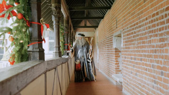 Personnage costumé en Léonard de Vinci marchant dans une galerie en briques du Château du Clos Lucé, décorée de guirlandes de sapin et de rubans rouges pour Noël.