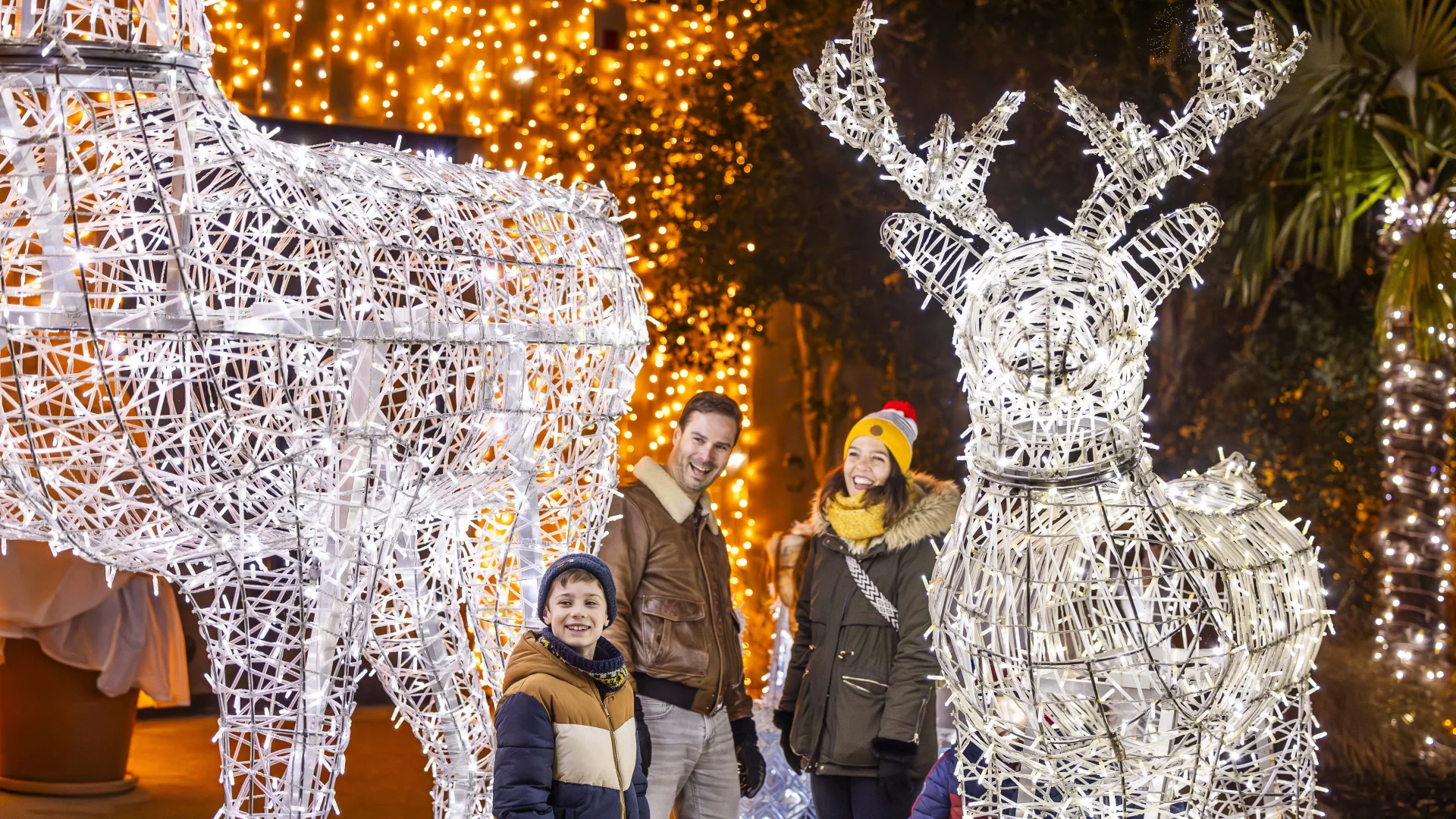 Une famille souriante composée d'un homme, d'une femme et d'un garçon pose le soir entre deux grandes sculptures lumineuses en forme de rennes réalisées avec des guirlandes électriques blanches. Des lumières dorées décorent les arbres et fondent une ambiance festive, rappelant les décorations de Noël.