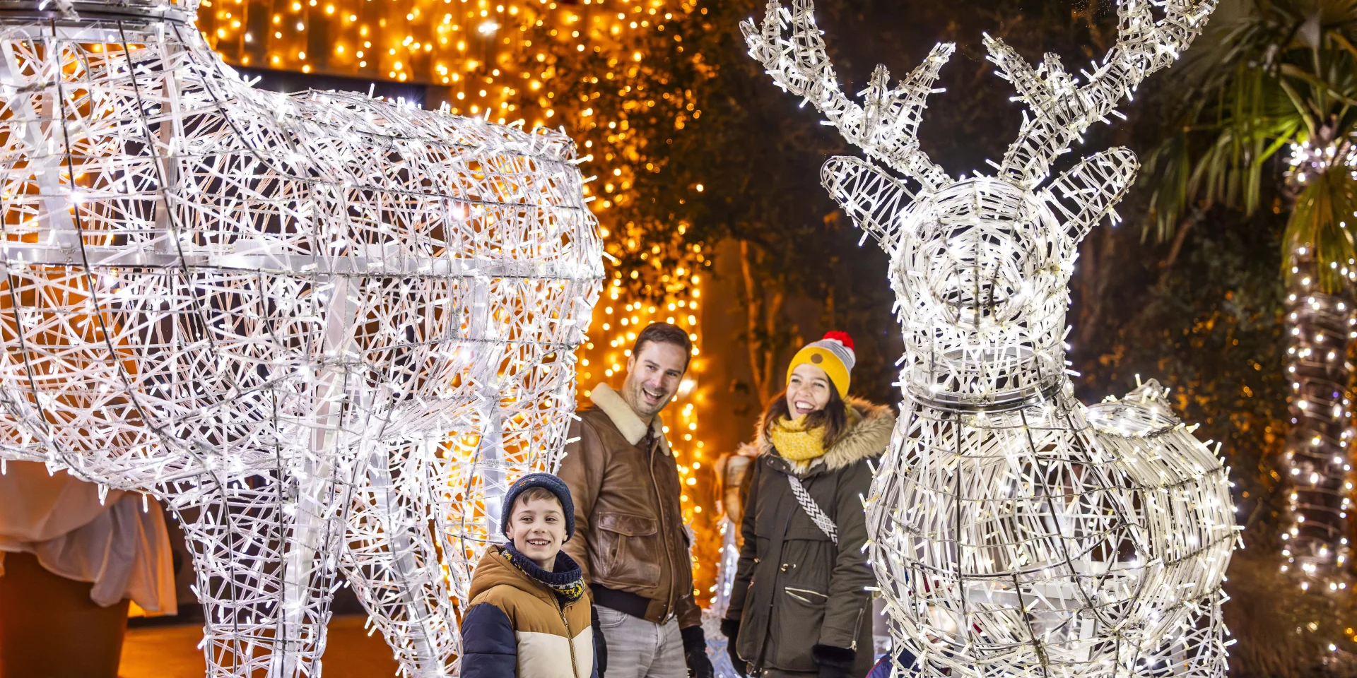 Une famille souriante composée d'un homme, d'une femme et d'un garçon pose le soir entre deux grandes sculptures lumineuses en forme de rennes réalisées avec des guirlandes électriques blanches. Des lumières dorées décorent les arbres et fondent une ambiance festive, rappelant les décorations de Noël.