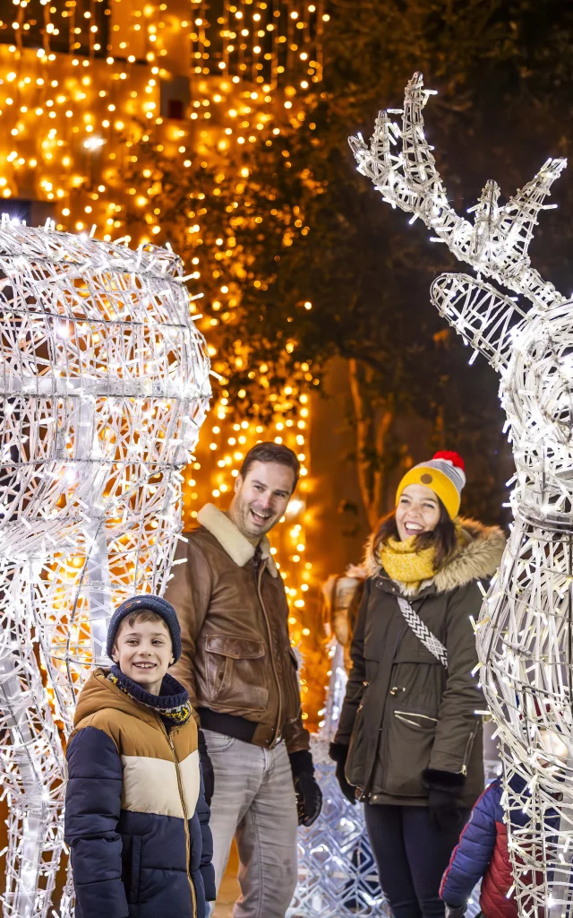 Une famille souriante composée d'un homme, d'une femme et d'un garçon pose le soir entre deux grandes sculptures lumineuses en forme de rennes réalisées avec des guirlandes électriques blanches. Des lumières dorées décorent les arbres et fondent une ambiance festive, rappelant les décorations de Noël.