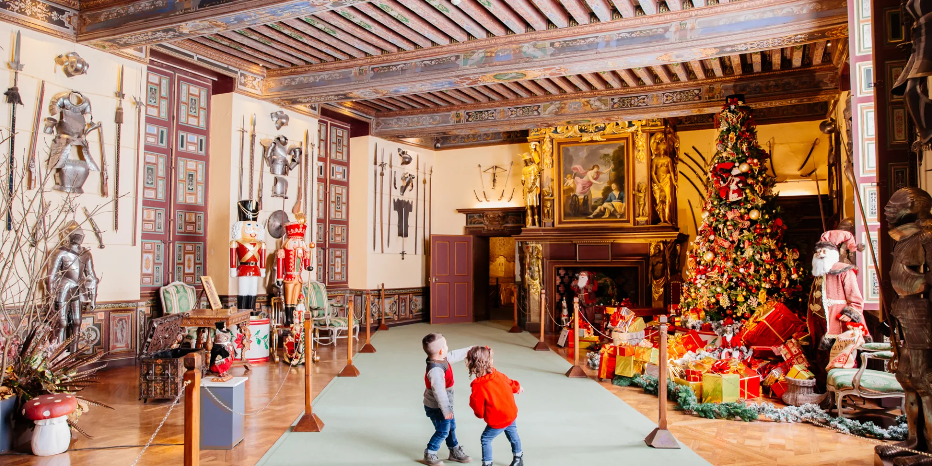 Deux enfants jouent dans une grande salle décorée de Noël au Château de Cheverny, entourée d’armures, de jouets géants et d’un sapin illuminé.