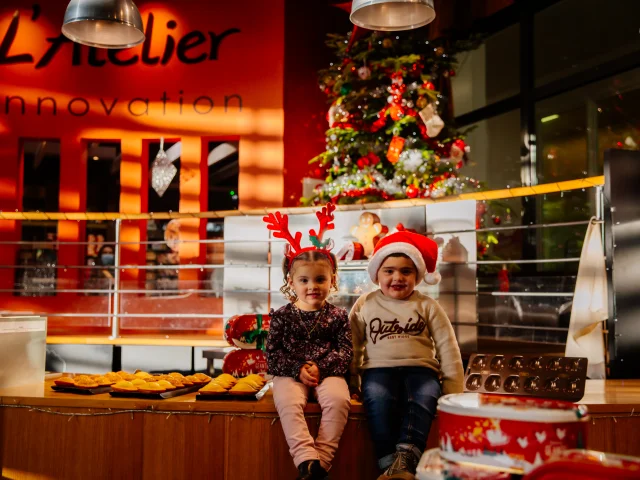 Deux jeunes enfants assis devant une table garnie de pâtisseries, dans un décor festif de Noël. L'un porte un serre-tête en forme de bois de renne et l'autre un bonnet de père Noël. Un sapin de Noël décoré se trouve à l'arrière-plan, ainsi que l'inscription L'Atelier Innovation sur un mur orange. L'ambiance est chaleureuse et festive.