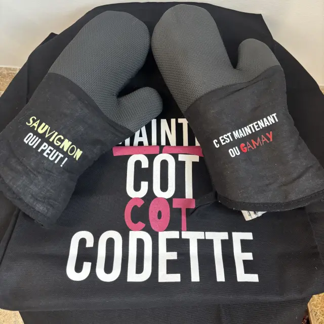 Deux gants de cuisine noirs avec jeux de mots sur les cépages, posés sur un tablier assorti.