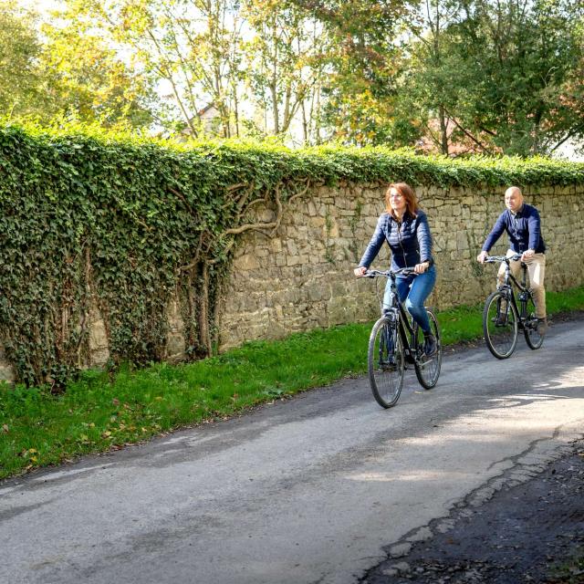 Une dame et un monsieur roulant à vélo sur un chemin asphalté le long d'un mur en pierre