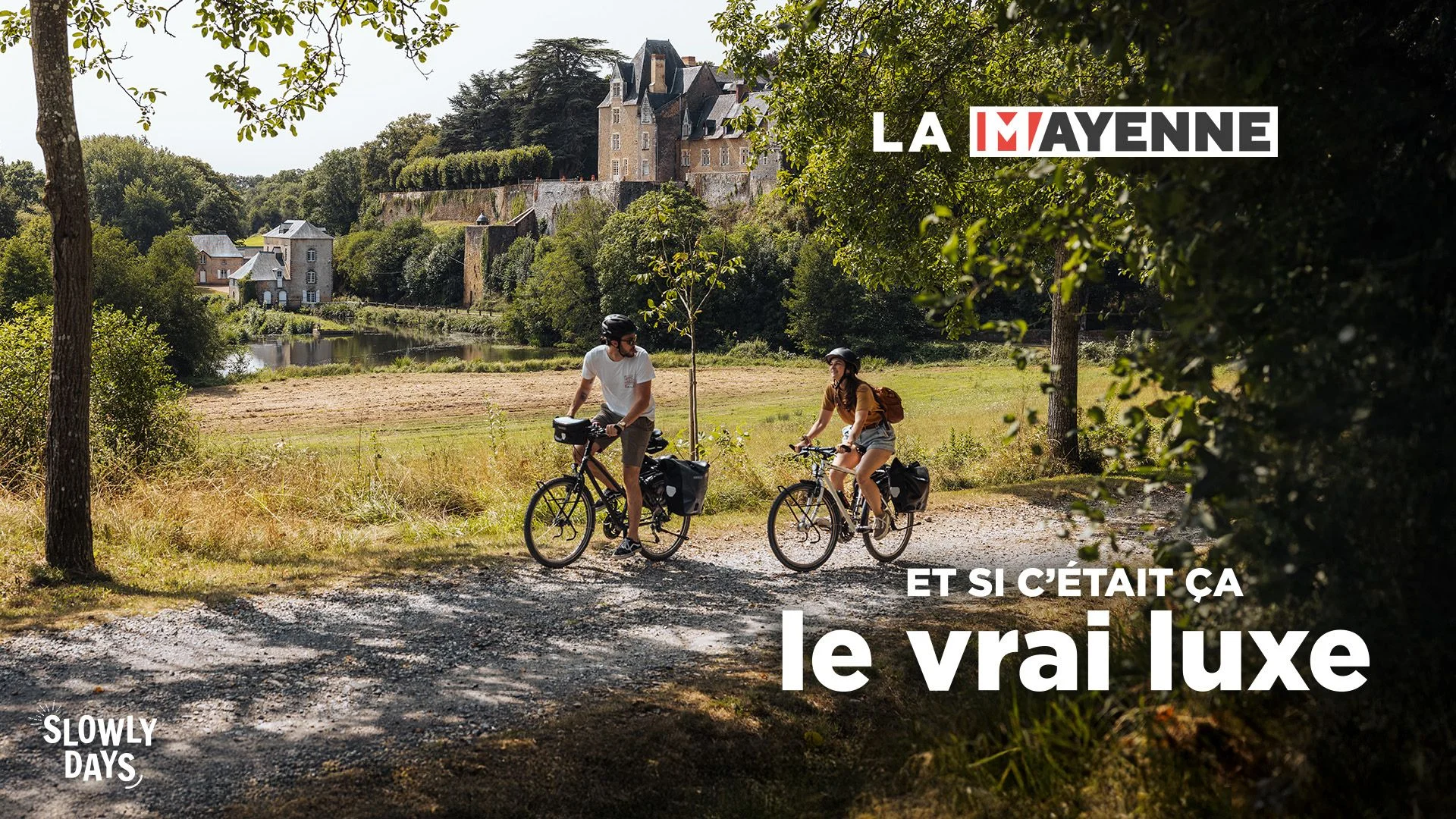 un homme et d'une femme en à vélo sur un chemin caillouteux ombragé. La scène se passe en été, tous deux portent short et tshirt, la végétation est dense et la luminosité intense. Leur vélo sont dotés de sacoches ce qui laissent penser que ce sont des cyclistes en itinérance. Le paysage est champêtre, ils roulent près d'un grand pré. En arrière plan, un château style renaissance domine une vallée où coule une rivière. Sur la berge on peut voir un moulin à eau. Dans le coin supérieur droit on voit la marque La Mayenne et dans le coin inférieur gauche le slogan Et si c'était ça le vrai luxe