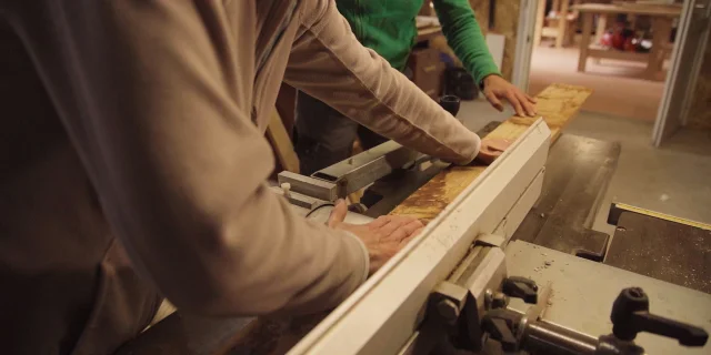 Deux personnes travaillent ensemble dans un atelier de menuiserie. L'une, vêtue d'un pull beige, guide une planche de bois clair à travers une machine-outil stationnaire. Ses mains sont concentrées sur la manipulation du bois. À côté, une autre personne portant un pull vert observe attentivement l'opération, sa main posée sur la planche pour la stabiliser. L'arrière-plan montre un espace de travail avec d'autres équipements et des éléments en bois. La scène suggère un travail manuel précis et collaboratif dans un environnement artisanal.