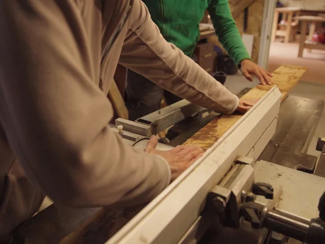 Deux personnes travaillent ensemble dans un atelier de menuiserie. L'une, vêtue d'un pull beige, guide une planche de bois clair à travers une machine-outil stationnaire. Ses mains sont concentrées sur la manipulation du bois. À côté, une autre personne portant un pull vert observe attentivement l'opération, sa main posée sur la planche pour la stabiliser. L'arrière-plan montre un espace de travail avec d'autres équipements et des éléments en bois. La scène suggère un travail manuel précis et collaboratif dans un environnement artisanal.