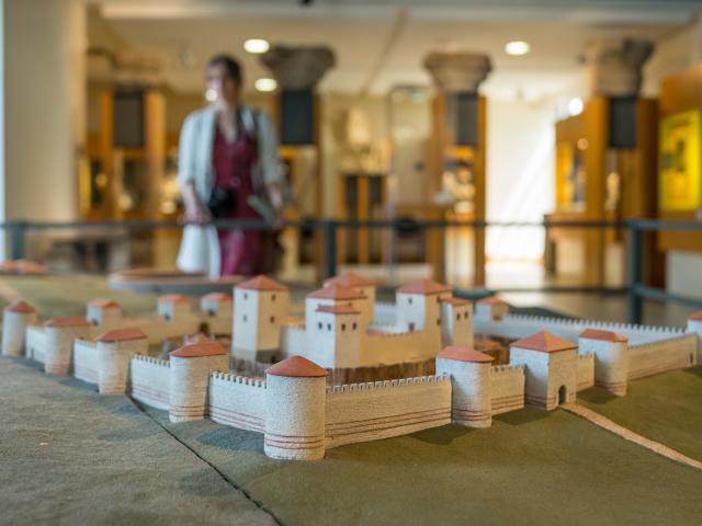 Photo prise à l'intérieur du musée archéologique départemental. Gros plan sur une maquette représentant la cité Gallo-Romaine antique de Noviodunum, aujourd'hui Jublains en Mayenne. L'arrière plan est flou mais on distingue une femme qui visite le musée.