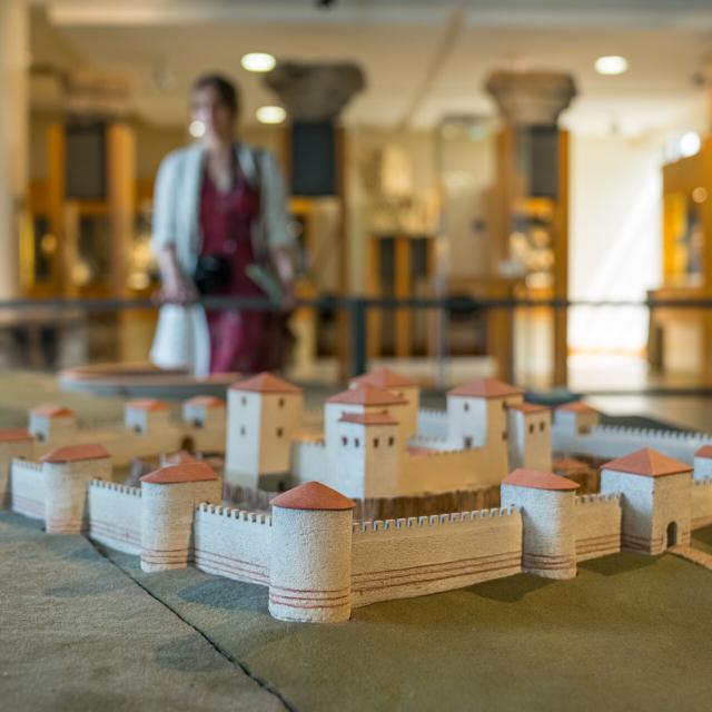 Photo prise à l'intérieur du musée archéologique départemental. Gros plan sur une maquette représentant la cité Gallo-Romaine antique de Noviodunum, aujourd'hui Jublains en Mayenne. L'arrière plan est flou mais on distingue une femme qui visite le musée.