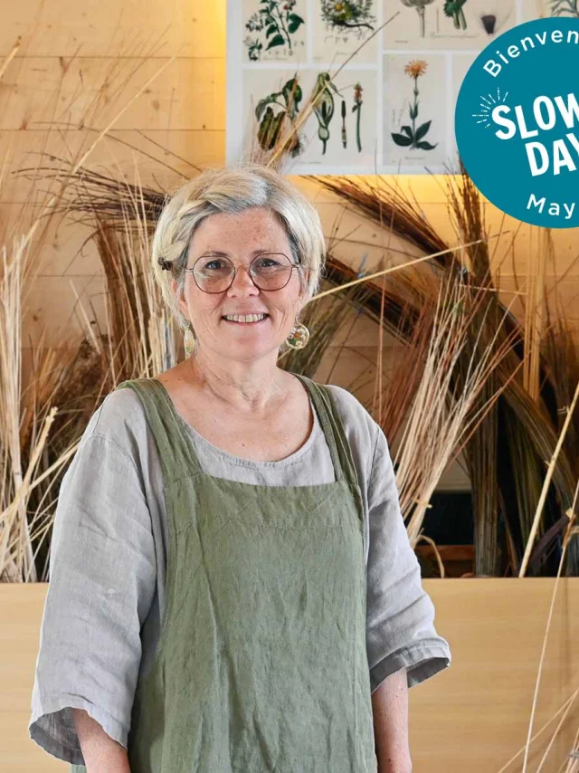Judith Charpentier de face dans son atelier. Derrierre elle, au mur on voit une affiche qui présente différents végétaux un peu comme les vieilles affiches des écoles d'antan. On voit également différentes tiges d'osier. Sur ce visuel en haut à droite on voit le logo Slowlydays