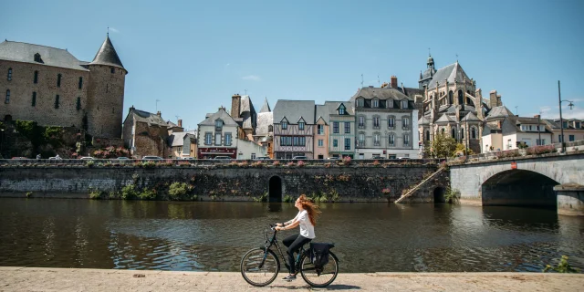 Une jeune femme rousse au cheveux long fait du vélo le long de la rivière la Mayenne sur un quai en plein centre ville de Mayenne par une journée très ensoleillée. Au deuxième plan on voit le bâti caractéristique de la ville avec des maisons à pan de bois ainsi que la basilique notre dame des miracle à droite et le château de Mayenne à gauche.