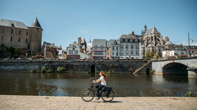 Une jeune femme rousse au cheveux long fait du vélo le long de la rivière la Mayenne sur un quai en plein centre ville de Mayenne par une journée très ensoleillée. Au deuxième plan on voit le bâti caractéristique de la ville avec des maisons à pan de bois ainsi que la basilique notre dame des miracle à droite et le château de Mayenne à gauche.