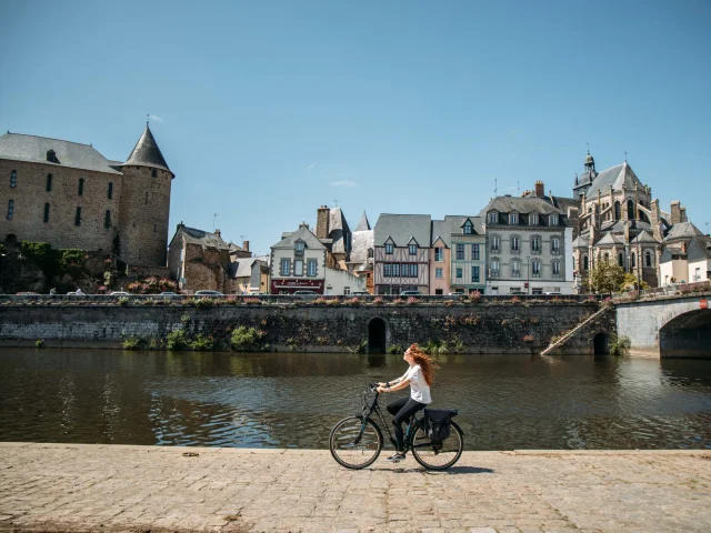 Une jeune femme rousse au cheveux long fait du vélo le long de la rivière la Mayenne sur un quai en plein centre ville de Mayenne par une journée très ensoleillée. Au deuxième plan on voit le bâti caractéristique de la ville avec des maisons à pan de bois ainsi que la basilique notre dame des miracle à droite et le château de Mayenne à gauche.