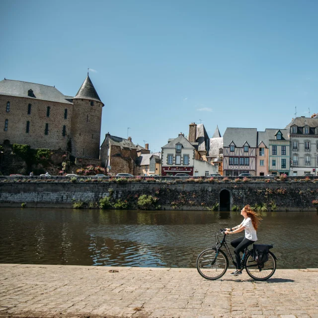 Une jeune femme rousse au cheveux long fait du vélo le long de la rivière la Mayenne sur un quai en plein centre ville de Mayenne par une journée très ensoleillée. Au deuxième plan on voit le bâti caractéristique de la ville avec des maisons à pan de bois ainsi que la basilique notre dame des miracle à droite et le château de Mayenne à gauche.