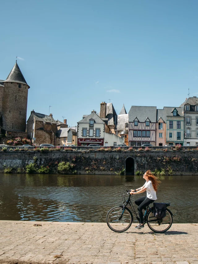 Une jeune femme rousse au cheveux long fait du vélo le long de la rivière la Mayenne sur un quai en plein centre ville de Mayenne par une journée très ensoleillée. Au deuxième plan on voit le bâti caractéristique de la ville avec des maisons à pan de bois ainsi que la basilique notre dame des miracle à droite et le château de Mayenne à gauche.