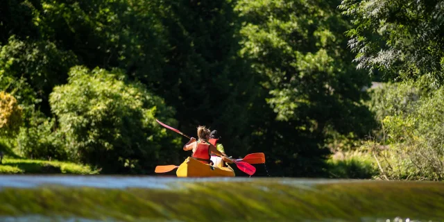 On voit deux personnes de dos naviguant sur un canoë. Les berges de la rivière sont arborées