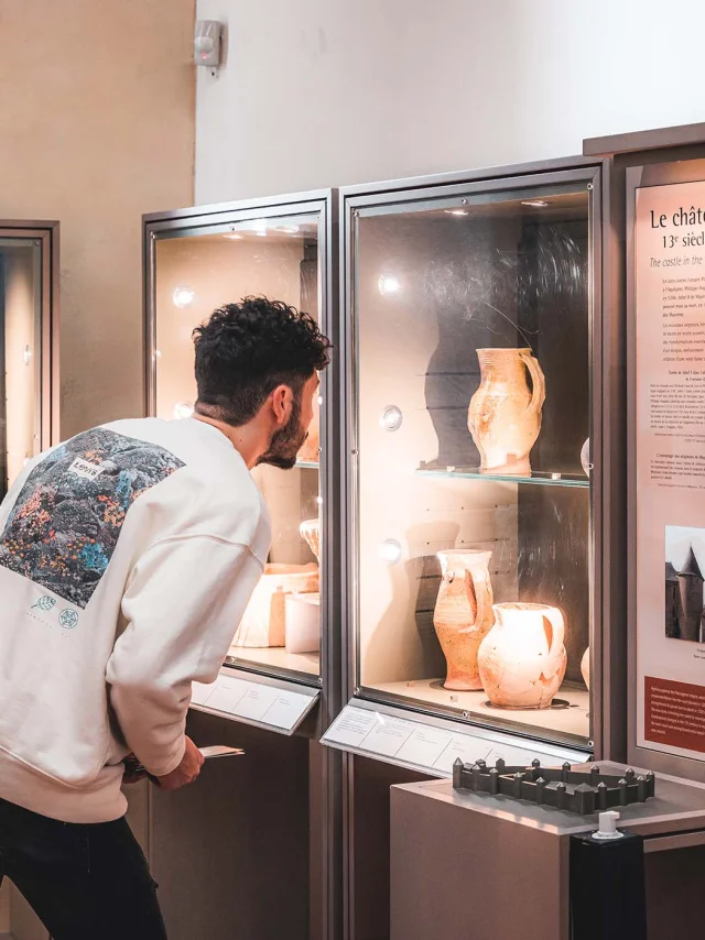 Au musée du château de Mayenne, un homme de trois quart dos observe une vitrine d'exposition dans laquelle sont exposées des poteries du Moyen Age. A côté de cette vitrine, on peut voir un panneau explicatif dont le titre est Le Château au treizième siècle et sur lequel on distingue différentes photos. Devant ce panneau, un maquette le l'enceinte du château est disposée, permettant aux personnes aveugles de la toucher pour avoir une idée de la disposition des lieux à l'époque médiévale.