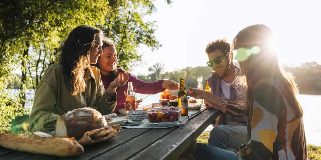 Groupe d’amis en pique-nique au bord d’un étang, partageant un repas au coucher du soleil.