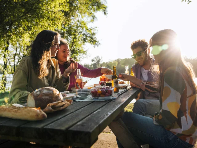 Groupe d’amis en pique-nique au bord d’un étang, partageant un repas au coucher du soleil.