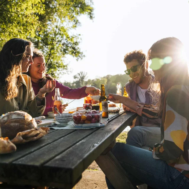 Groupe d’amis en pique-nique au bord d’un étang, partageant un repas au coucher du soleil.