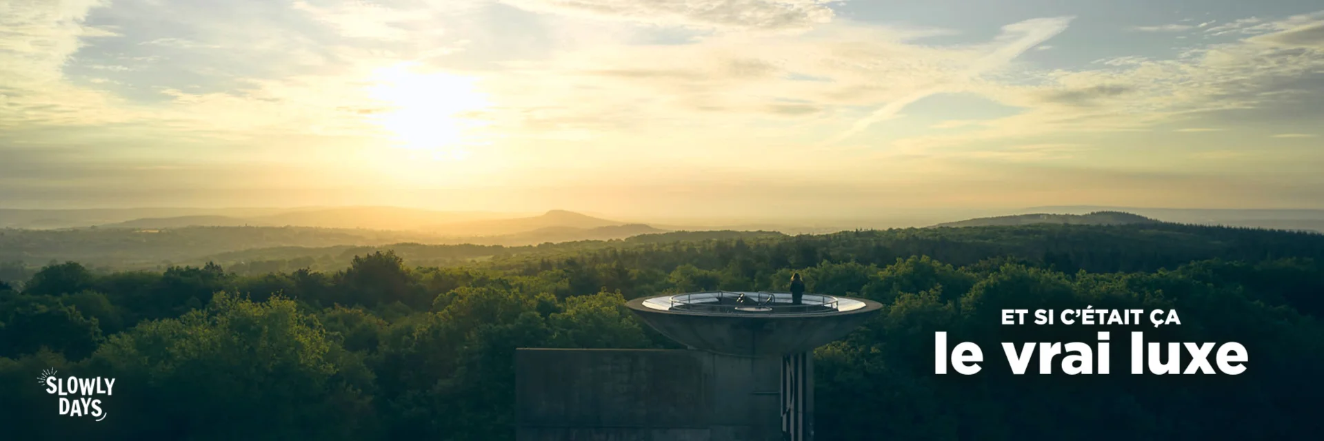 Visuel au format Paysage. Vue panoramique au coucher du soleil depuis le Belvédère du Mont des Avaloirs en Mayenne. On y voit une plateforme circulaire dominant une vaste forêt verdoyante sous un ciel doré. On distingue une silhouette sur la plateforme. Dans le coin supérieur droit on voit la marque La Mayenne et dans le coin inférieur droit le slogan Et si c'était ça le vrai luxe.