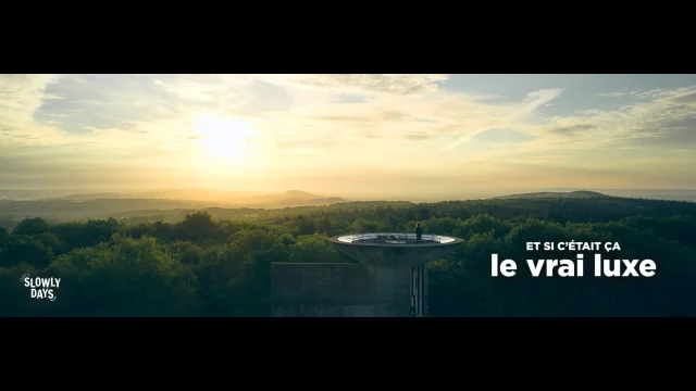Visuel au format Paysage. Vue panoramique au coucher du soleil depuis le Belvédère du Mont des Avaloirs en Mayenne. On y voit une plateforme circulaire dominant une vaste forêt verdoyante sous un ciel doré. On distingue une silhouette sur la plateforme. Dans le coin supérieur droit on voit la marque La Mayenne et dans le coin inférieur droit le slogan Et si c'était ça le vrai luxe.