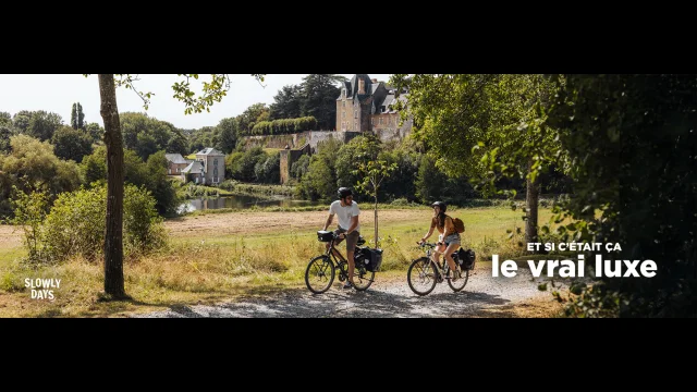 Visuel au format Paysage. Un homme et d'une femme en à vélo sur un chemin caillouteux ombragé. La scène se passe en été, tous deux portent short et tshirt, la végétation est dense et la luminosité intense. Leur vélo sont dotés de sacoches ce qui laissent penser que ce sont des cyclistes en itinérance. Le paysage est champêtre, ils roulent près d'un grand pré. En arrière plan, un château style renaissance domine une vallée où coule une rivière. Sur la berge on peut voir un moulin à eau. Dans le coin supérieur droit on voit la marque La Mayenne et dans le coin inférieur gauche le slogan Et si c'était ça le vrai luxe