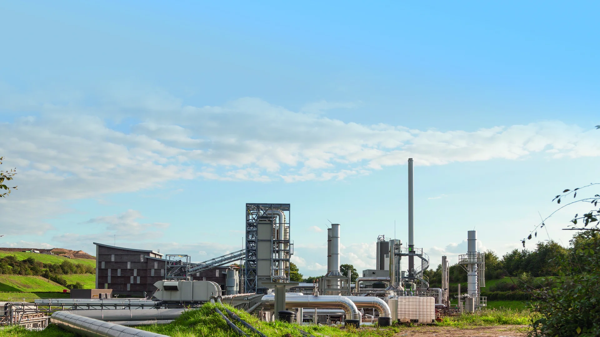 Vue de jour d'une usine de traitement de déchets Seche Environnement avec des tuyaux, des cheminées et un bâtiment en bois, sur un site industriel entouré de végétation et de collines.