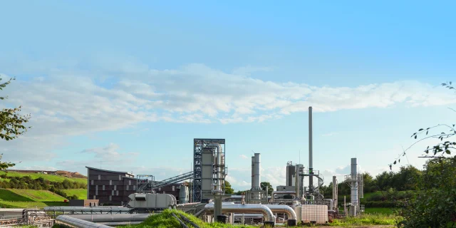Vue de jour d'une usine de traitement de déchets Seche Environnement avec des tuyaux, des cheminées et un bâtiment en bois, sur un site industriel entouré de végétation et de collines.