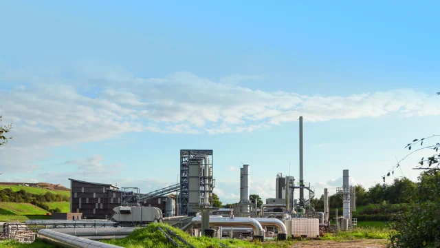 Vue de jour d'une usine de traitement de déchets Seche Environnement avec des tuyaux, des cheminées et un bâtiment en bois, sur un site industriel entouré de végétation et de collines.