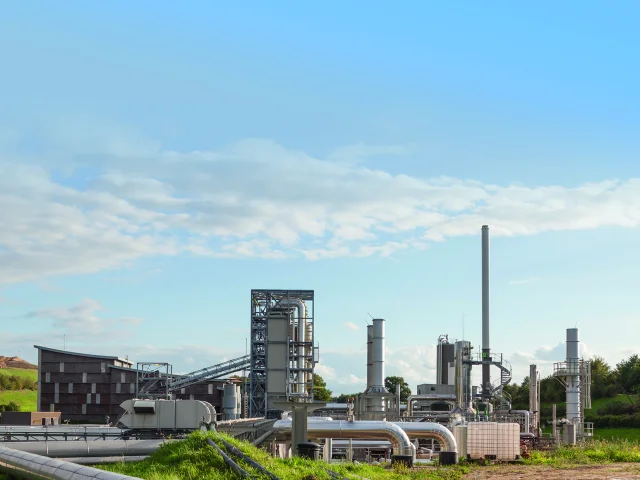 Vue de jour d'une usine de traitement de déchets Seche Environnement avec des tuyaux, des cheminées et un bâtiment en bois, sur un site industriel entouré de végétation et de collines.