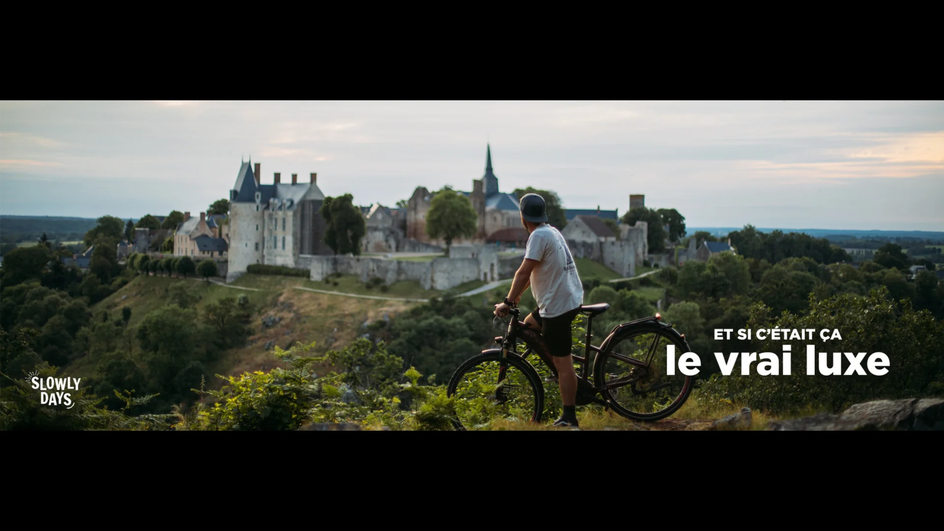 Visuel au format Paysage. Un homme sur un VTT fait une pause au Tertre Ganne qui est un promontoire en pleine nature ayant une superbe vue sur le village de Sainte-Suzanne. Ce village est perché sur une hauteur, on voit très bien le château et les remparts. L'homme porte un short, un tshirt et une casquette, on est en été, en fin de journée, la luminosité est assez douce, on s'approche du coucher de soleil. Dans le coin supérieur droit on voit la marque La Mayenne et dans le coin inférieur droit le slogan Et si c'était ça le vrai luxe