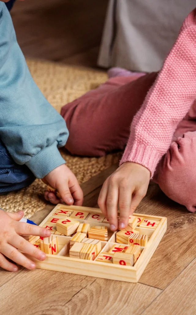 Deux enfants assis par terre jouant avec des lettres en bois dans un plateau éducatif.
