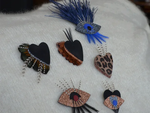 Collection de broches artisanales en forme de cœurs et d'yeux, ornées de plumes et de paillettes, posées sur un tissu blanc.