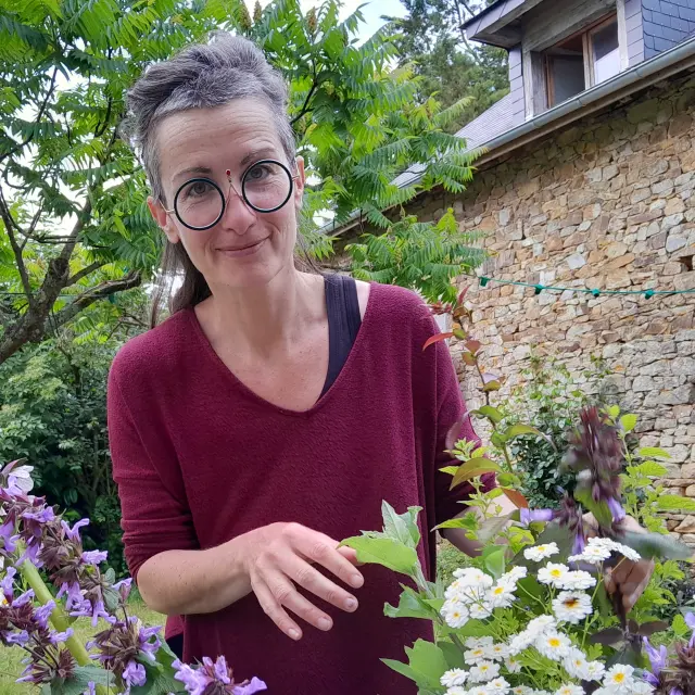 Portrait de Julie Pinçon dans son jardin. Au premier plan on voit des bouquets qu'elle confectionne. En arrière plan on duistingue une maison en pierre de taille.